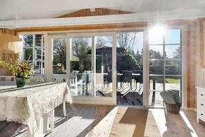 A room with a sliding glass door leading to an outdoor deck, a table with a white tablecloth, and a wicker basket with cushions. - Lovely cottage in Hals 250 m to the beach (Hals)