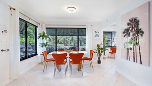 Un espace repas moderne avec une table ronde, six chaises orange et de grandes fenêtres équipées de stores noirs.