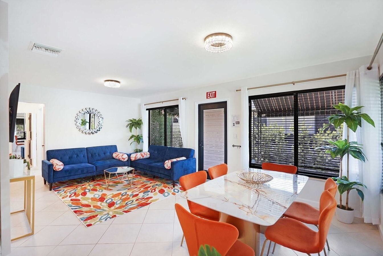 Ein modernes Wohnzimmer mit einem blauen Sofa, orangefarbenen Sesseln, einem Esstisch und einem farbenfrohen Teppich.
