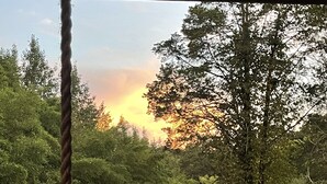 A wooden deck with a table and chairs, overlooking a forested area with a sunset in the background. - Craftsman style family gathering home close to Guntersville (Scottsboro)