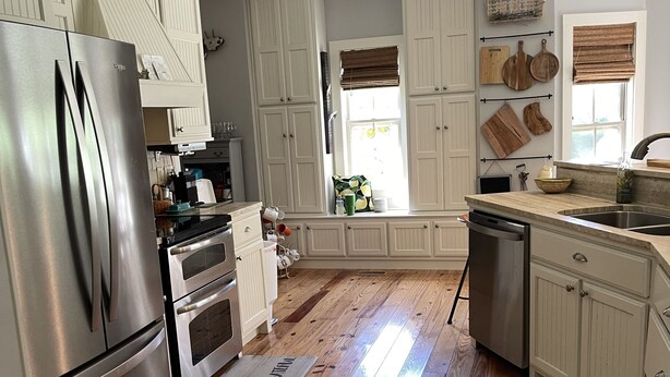 A kitchen with wooden flooring, white cabinetry, a stainless steel refrigerator, and a window with blinds. - Craftsman style family gathering home close to Guntersville (Scottsboro)