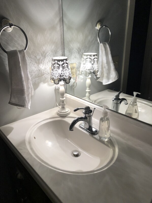 Une salle de bain avec un lavabo blanc, un miroir, un porte-serviette, une lampe et un distributeur de savon.