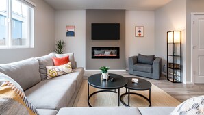 A modern living room with a grey sofa, a grey armchair, a fireplace, a flat-screen TV, and a floor lamp.