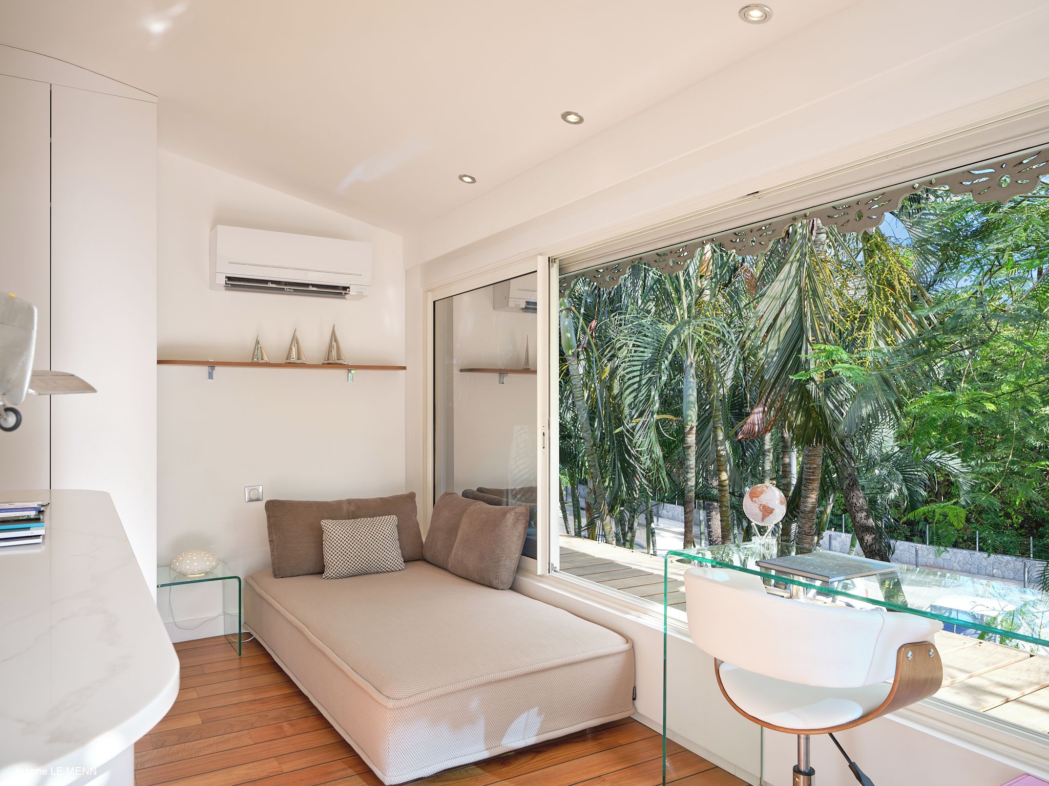 A modern bedroom with a sofa bed, a glass table, and a large window with a view of tropical plants.