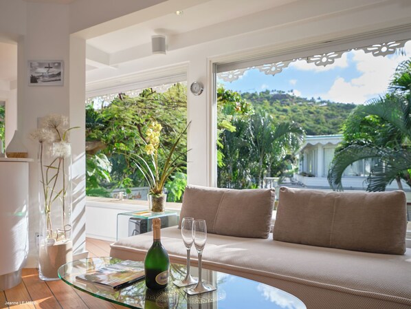 Una sala de estar moderna con una mesa de centro de vidrio, un sofá y vistas a una vegetación exuberante y una ladera.