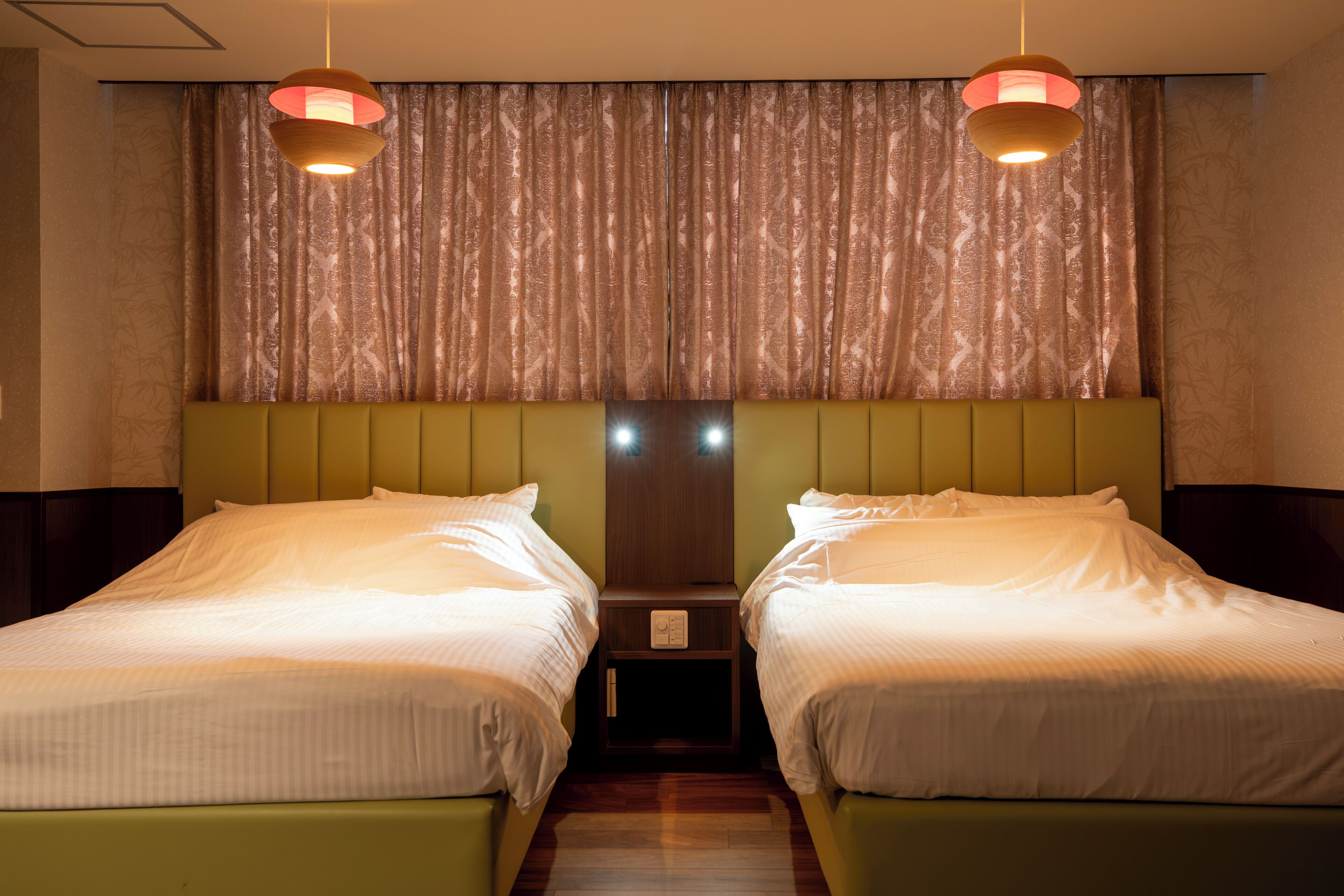 Une chambre d’hôtel avec deux lits simples, une tête de lit en bois et un rideau orné d’un motif de feuilles.