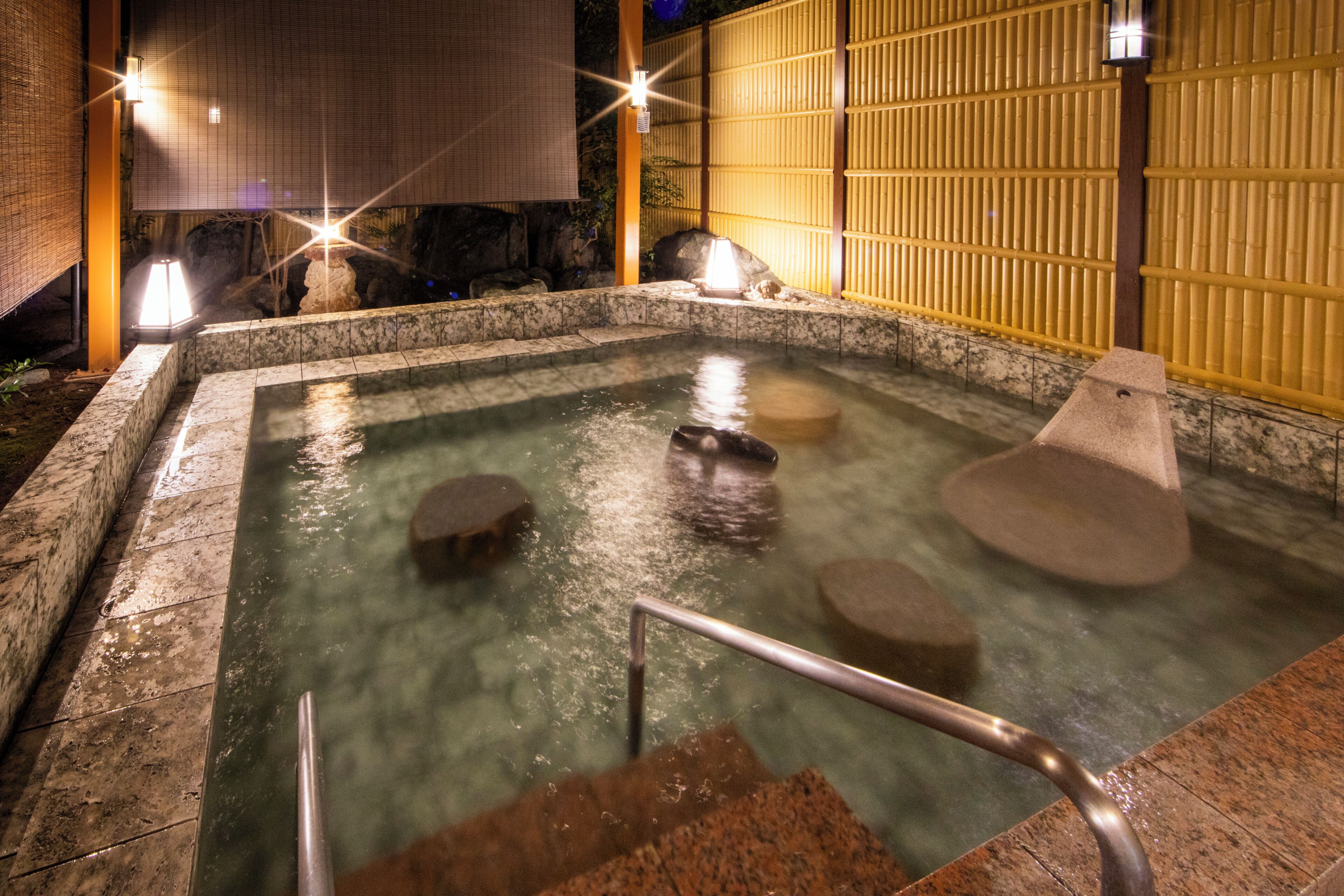 Un bain thermal en plein air, avec des marches en pierre menant à l’eau, entouré d’une clôture en bambou et de murs de pierre.