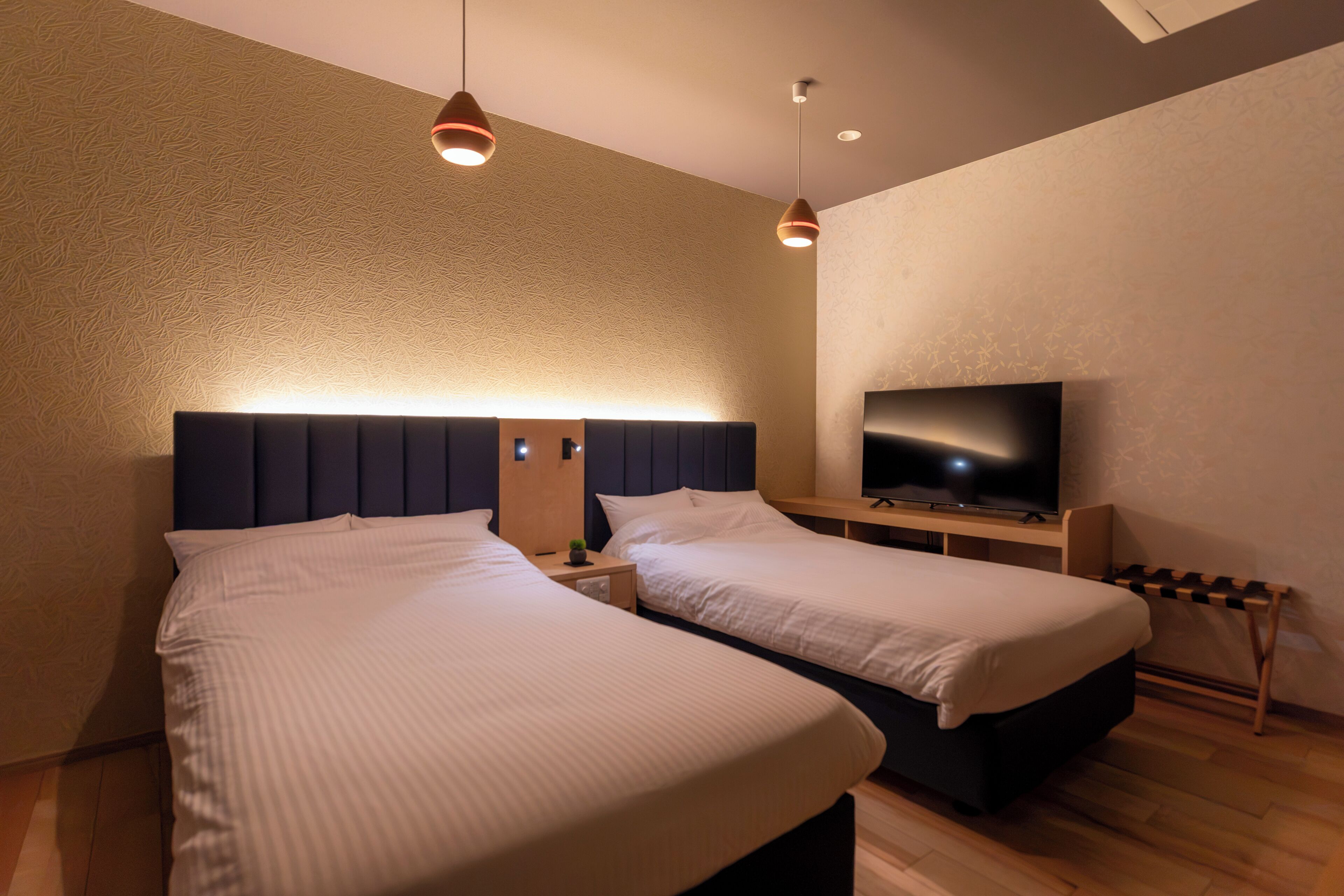 A modern hotel room with two beds, a flat-screen TV, and pendant lighting.