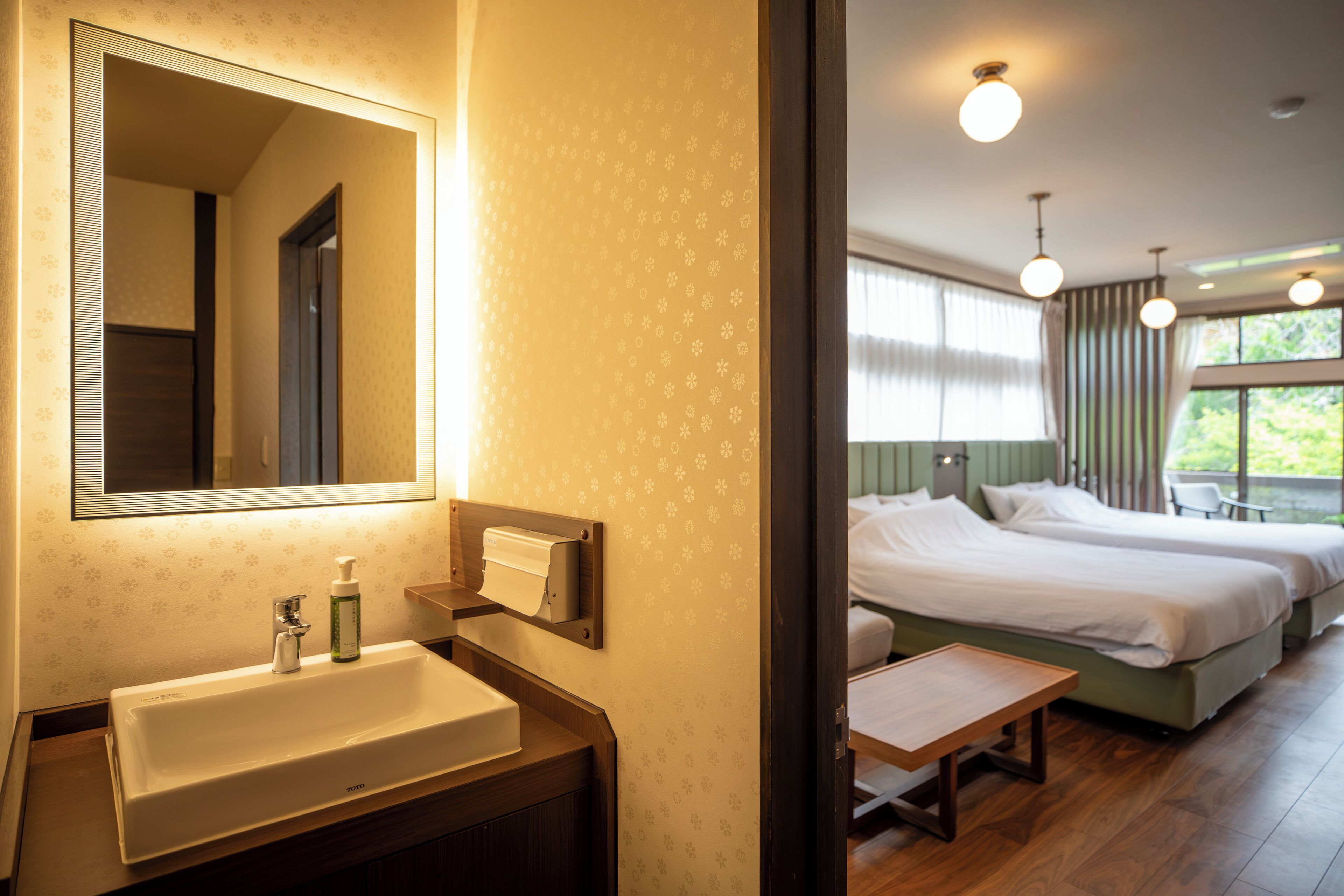Een hotelkamer met een grote spiegel, een wastafel, een nachtkastje, twee bedden en een raam met gordijnen.