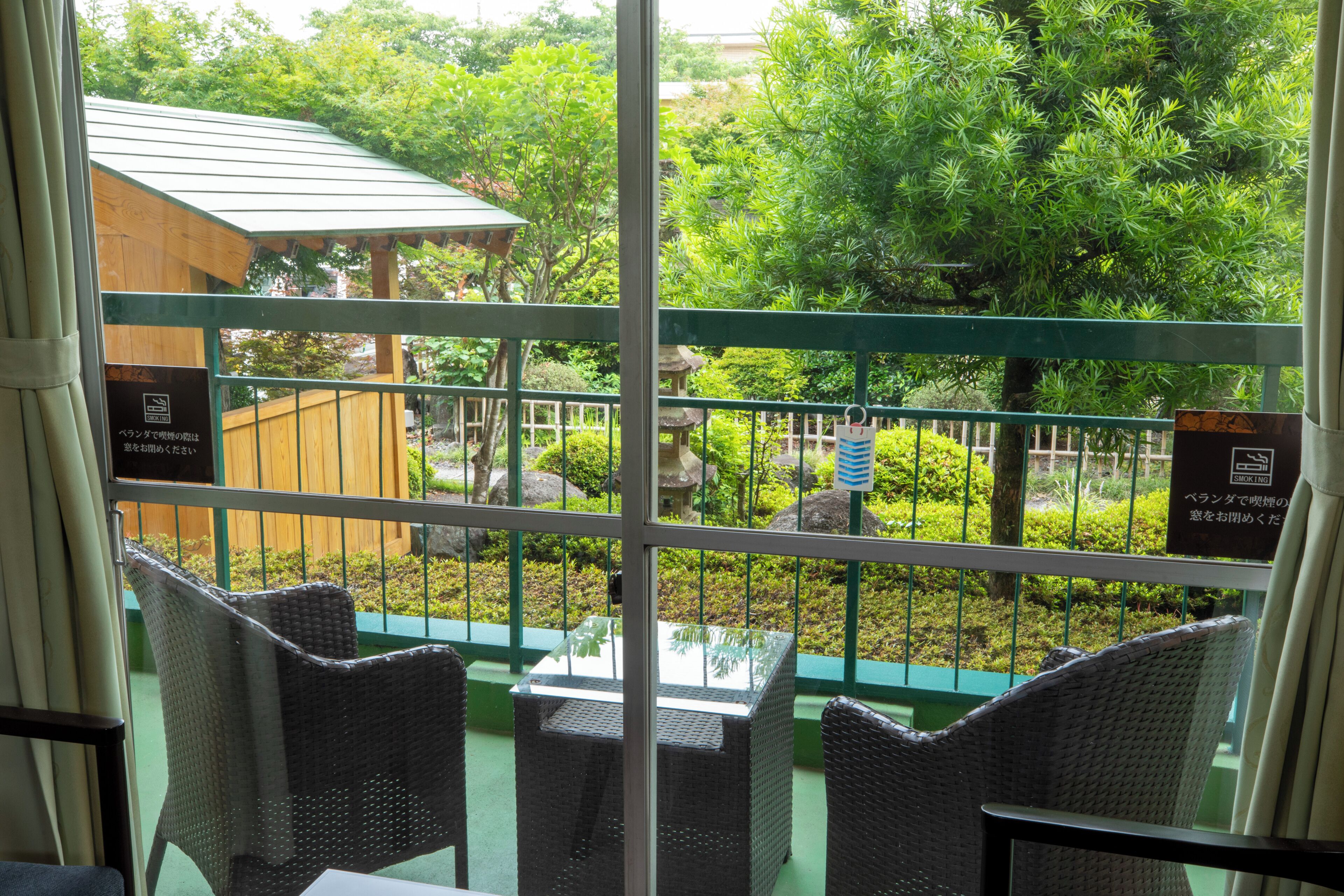 Ein Balkon mit Korbstühlen und einem Tisch, mit Blick auf einen Garten mit einer Holzkonstruktion und einem Schild, das einen Raucherbereich kennzeichnet.