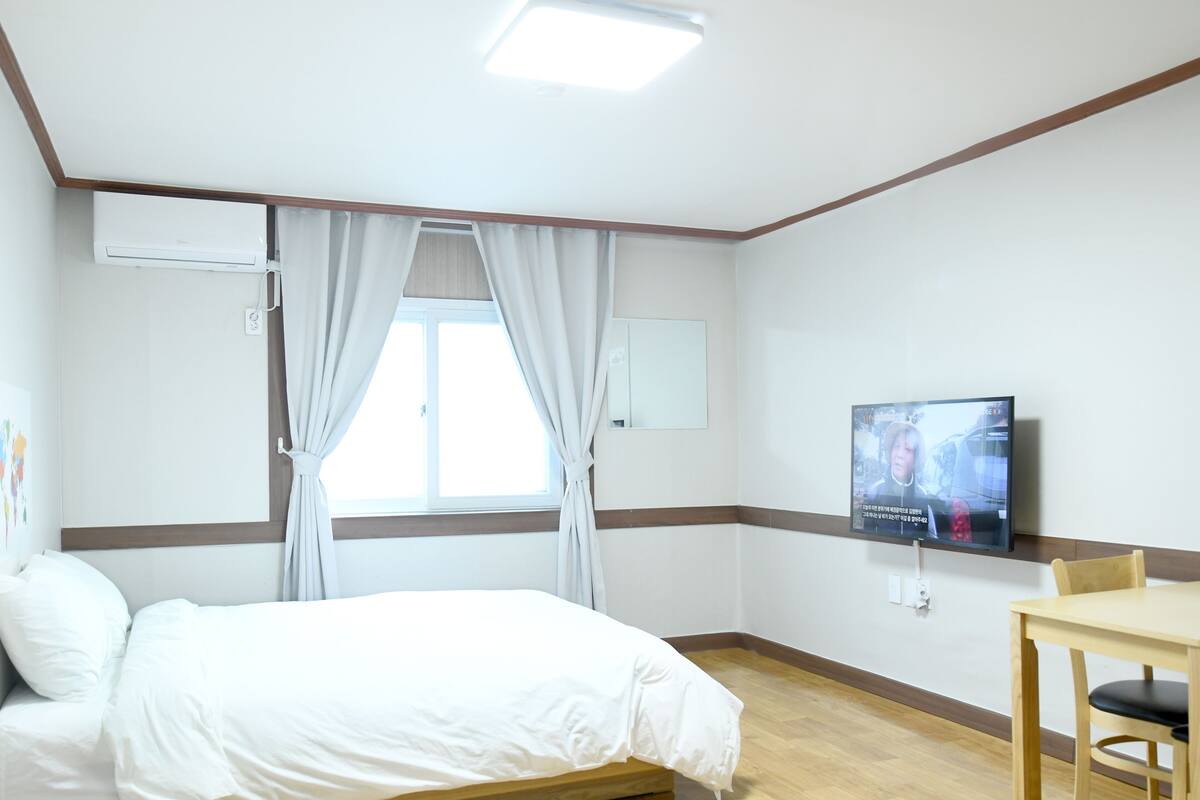 A hotel room with a bed, a TV, a desk, and a window with curtains.