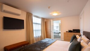 Habitación de hotel con una cama, un banco, un televisor, un baño con lavamanos y espejo, y una puerta que da a otra habitación.