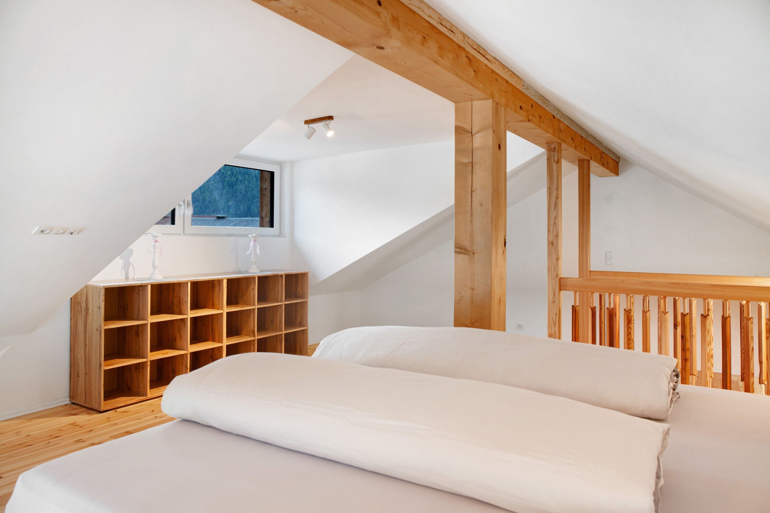Une chambre avec un plafond en pente, des poutres apparentes, une étagère intégrée en bois, un lit avec du linge de lit blanc et une fenêtre offrant une vue.