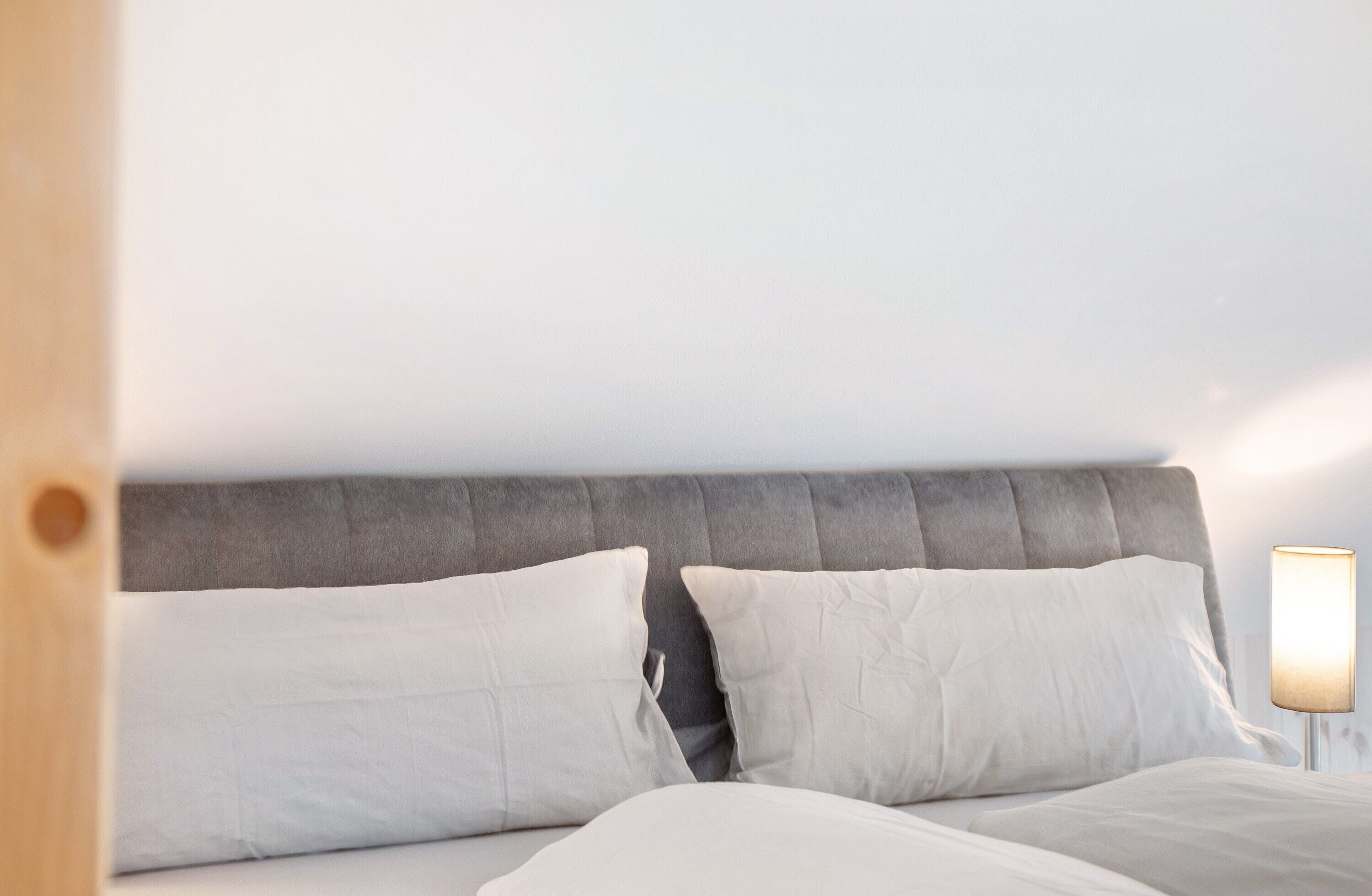 Une chambre à coucher avec une tête de lit rembourrée de couleur grise, du linge de lit blanc et une lampe de chevet.