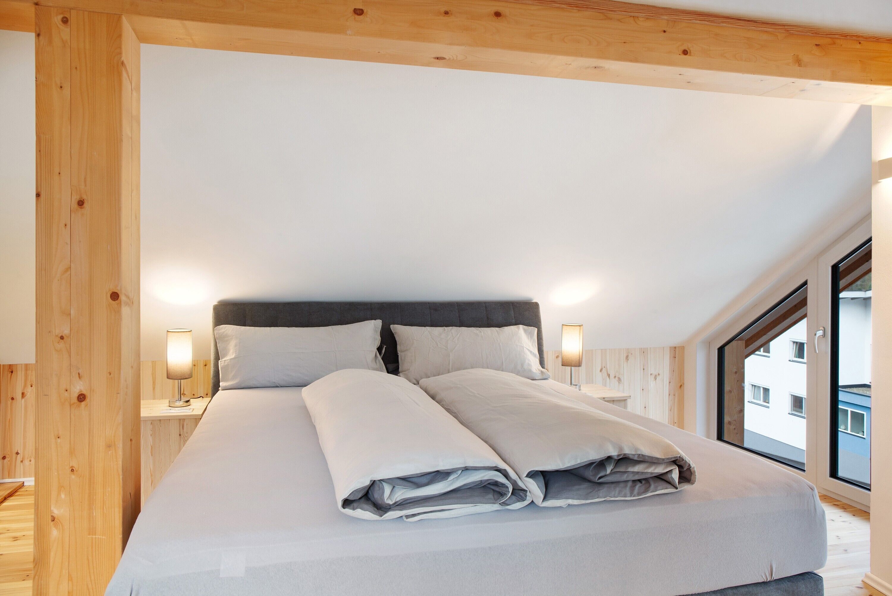 Un dormitorio con una cama grande, vigas de madera, una mesita de noche con lámpara y una ventana que ofrece vistas a edificios.