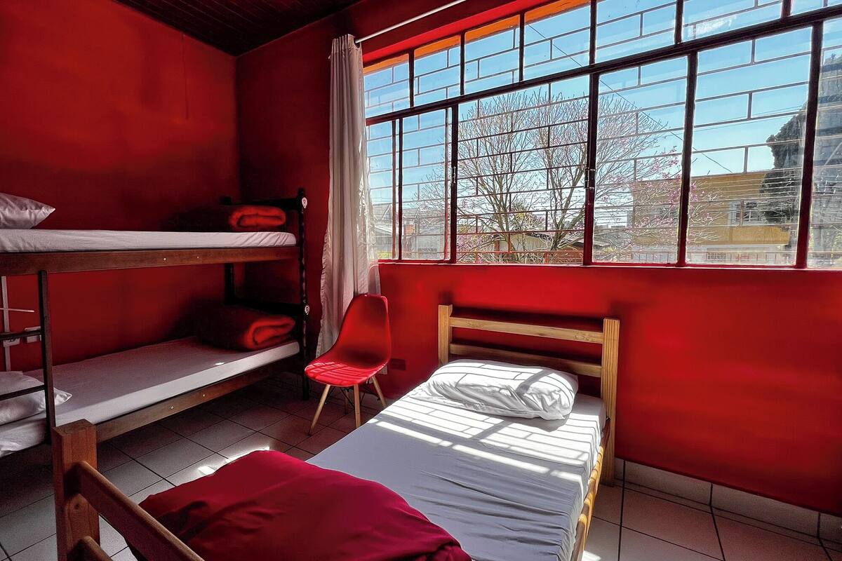 Habitación tipo dormitorio con literas, una cama individual, una silla roja y un ventanal con vistas a árboles y edificios.