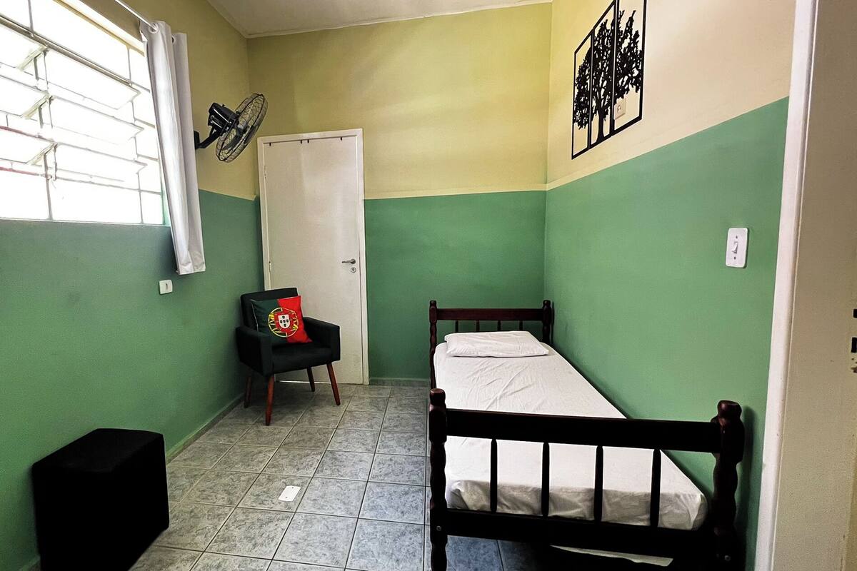 Una habitación pequeña con una cama individual, paredes verdes, una ventana con persianas, una silla con cojín de bandera y un ventilador en la pared.