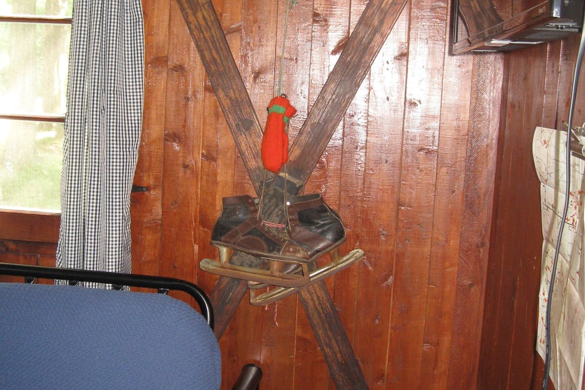 A room with wooden walls, a bed, a window with a curtain, and a wooden rocking chair with a red object on it.