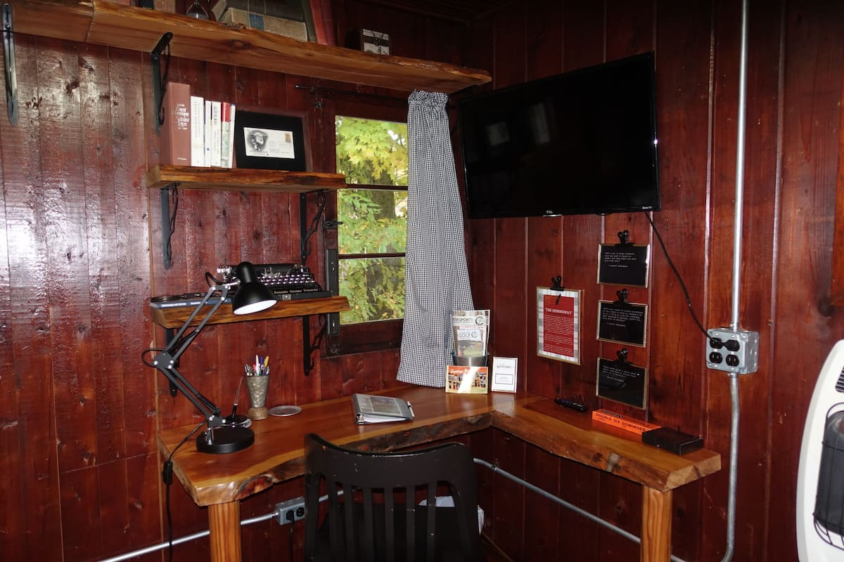 A wooden cabin with a desk, a computer, and a TV. There are shelves with books and a window with a view of trees.