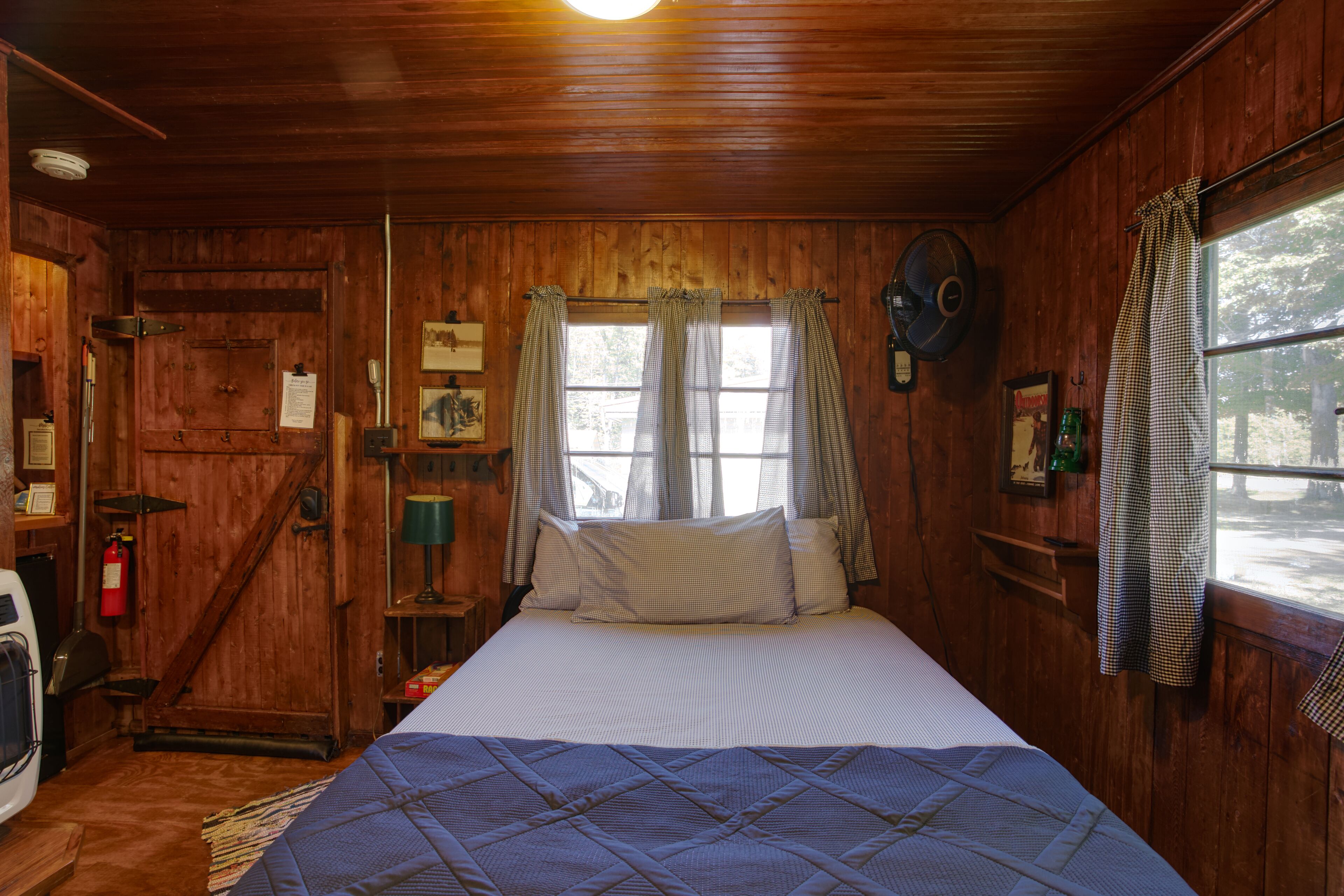 Une pièce en bois avec un lit, une fenêtre avec des rideaux, un ventilateur et un extincteur.