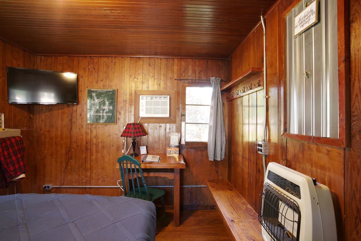 A wooden cabin room with a bed, desk, chair, television, and a heater.