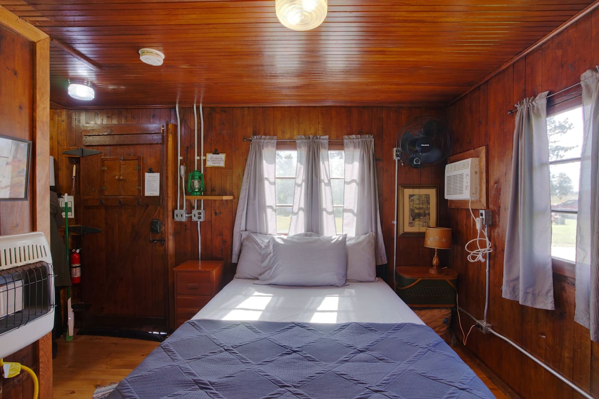 A wooden cabin room with a bed, a small table, a fan, and a window with curtains.