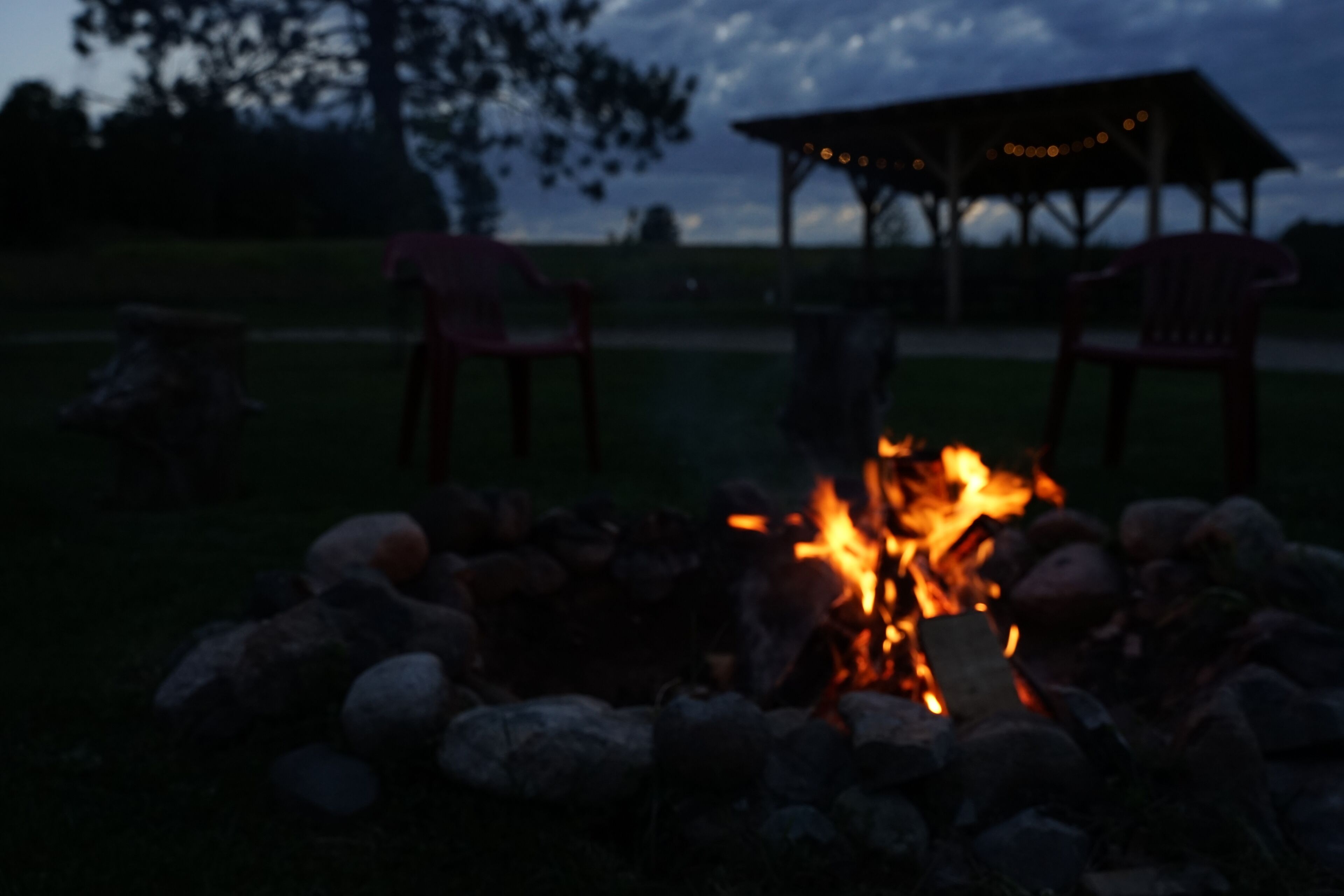 A campfire surrounded by rocks, with two red chairs and a gazebo in the background.