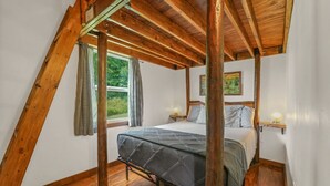 Une chambre avec un plafond en pente, des poutres en bois, un lit recouvert d’une courtepointe, deux tables de chevet avec des lampes et une fenêtre avec des rideaux.