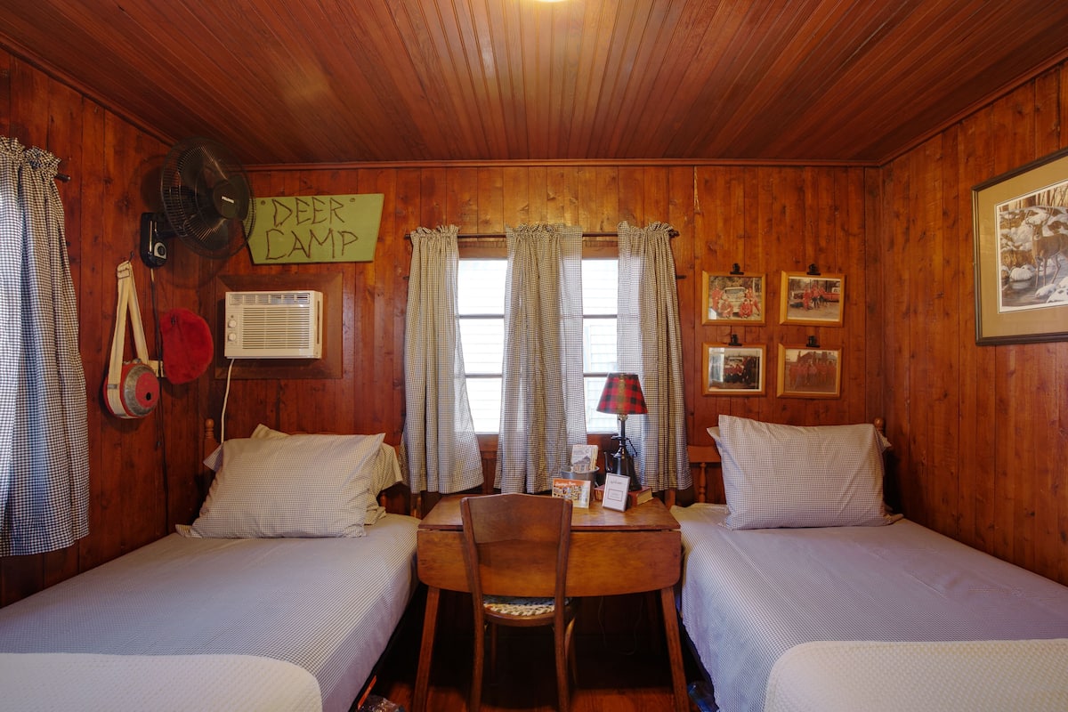 A cabin with two beds, a wooden table, a chair, a fan, a sign, and a window with curtains.