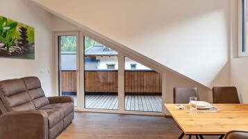 Ein Zimmer mit einem Holztisch, einem braunen Sofa und einem großen Fenster mit Blick auf eine Holzterrasse und ein Haus.