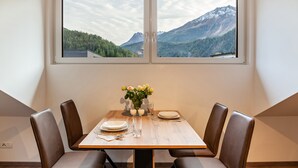 Comedor con una mesa preparada para dos personas, dos sillas, un florero con flores y vistas a la montaña a través de una ventana.