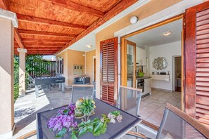 Un espace extérieur aménagé et abrité, avec une table et des chaises, une terrasse offrant une vue sur le jardin et une porte-fenêtre coulissante donnant sur la cuisine.