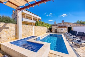 A swimming pool with a stone wall and lounge chairs, a covered area with a pergola, and a building with balconies.