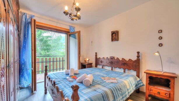 A bedroom with a wooden bed, a nightstand, a chandelier, a large window with curtains, and a view of greenery outside. - Appartamento 7 Bello Mountain View, Sauze d'Oulx, Italy (Sauze d'Oulx)