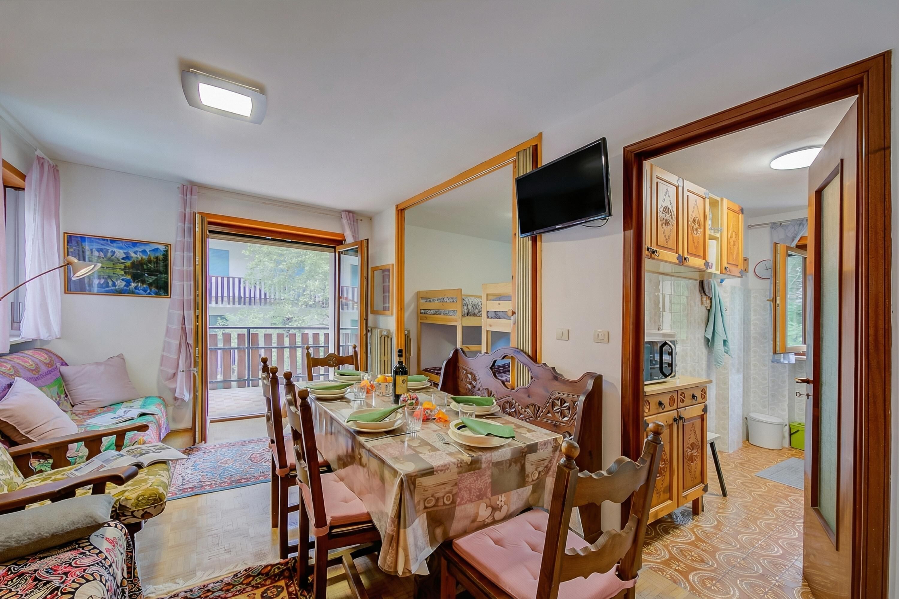 Un coin repas avec une table dressée pour quatre personnes, des chaises en bois, un téléviseur fixé au mur et une vue sur le balcon extérieur.