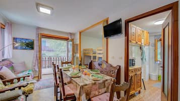 Un coin repas avec une table dressée pour quatre personnes, des chaises en bois, un téléviseur fixé au mur et une vue sur le balcon extérieur.