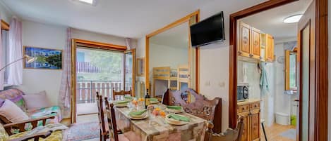 A dining area with a table set for four, wooden chairs, a television mounted on the wall, and a view of the outside balcony.