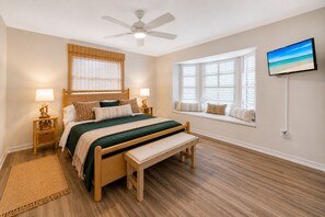 Une chambre à coucher comprenant un lit, des tables de chevet, un banc, un ventilateur de plafond, une fenêtre avec des stores et un téléviseur fixé au mur.
