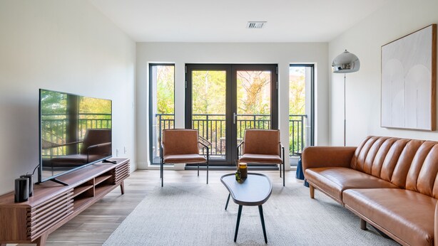 Una sala de estar moderna con televisor, sofá de cuero y sillones, ventanas amplias y un balcón.
