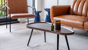 Una sala de estar moderna con un sofá de cuero marrón, un sillón a juego, una mesa de centro y una lámpara de pie. Hay un ventanal con vistas a árboles y un balcón.