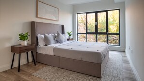 Un dormitorio moderno con una cama grande, una mesita de noche, una planta en maceta y una ventana con vista a árboles.