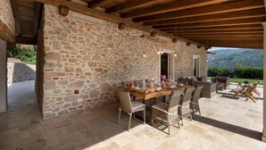Zona pranzo all'aperto con pareti in pietra, un tavolo di legno e sedie in vimini, con vista su un paesaggio montuoso.