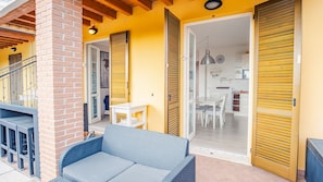 Une pièce avec un canapé en osier bleu, une table à manger avec des chaises, et un coin cuisine visible à travers une porte ouverte.