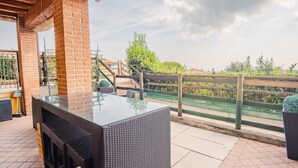 Un patio al aire libre con pared de ladrillo, muebles de mimbre y vista a un jardín y edificios al fondo.