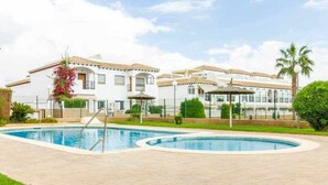 A swimming pool with a pergola and palm trees, surrounded by a white building with a tiled roof. - Bungalow Pink Punta Prima (Punta Prima)