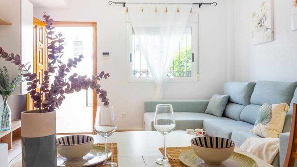 A modern living room with a sofa, a dining table with bowls and wine glasses, and a window with sheer curtains. - Bungalow Pink Punta Prima (Punta Prima)