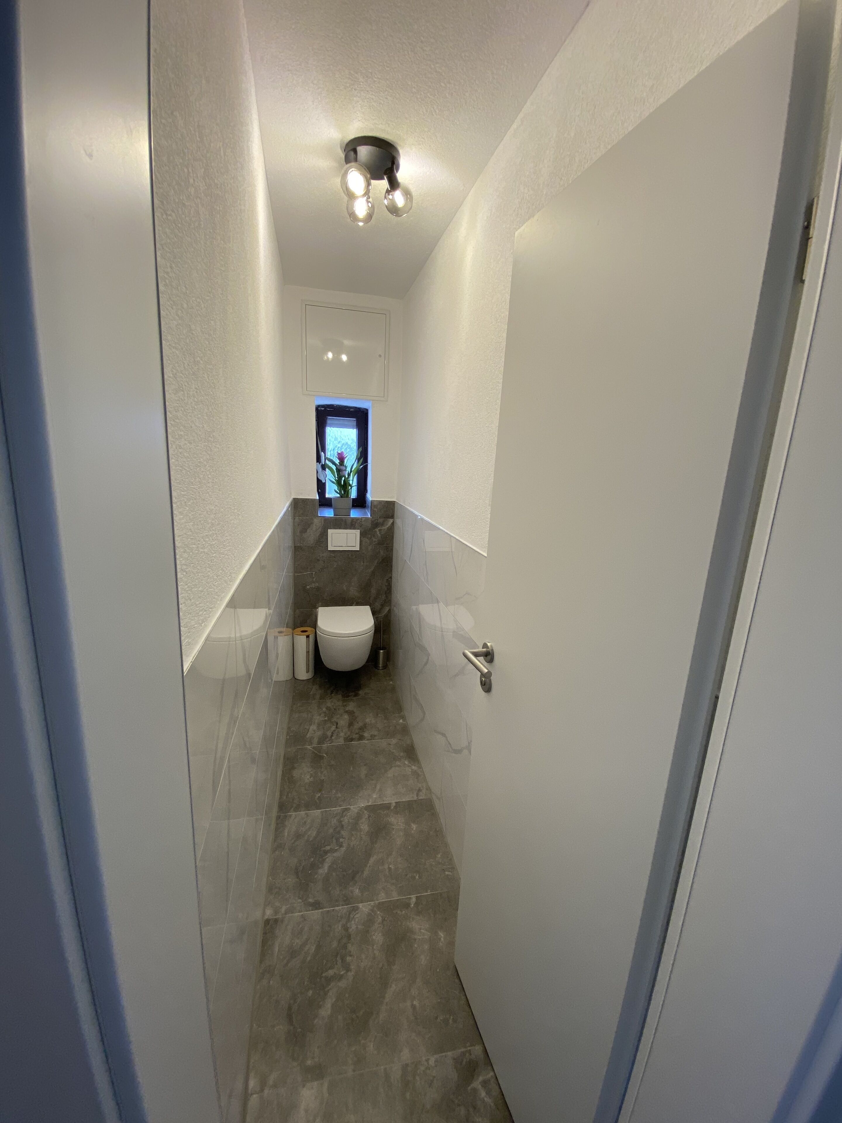 Un couloir étroit mène à une salle de bain équipée de toilettes, d’une petite fenêtre et d’un luminaire au plafond.