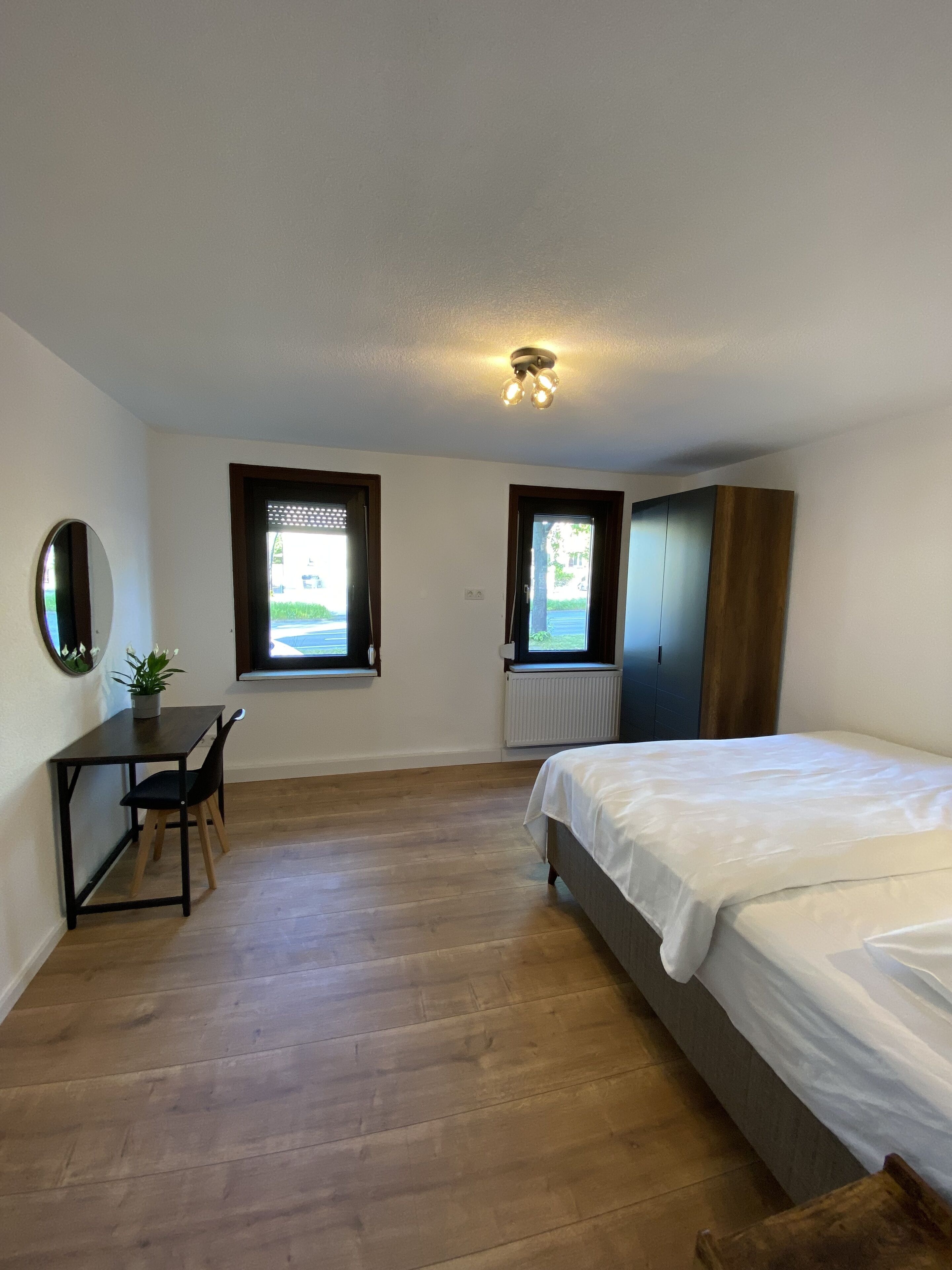 Ein Schlafzimmer mit Bett, Schreibtisch, Stuhl, zwei Fenstern und einer Heizung.