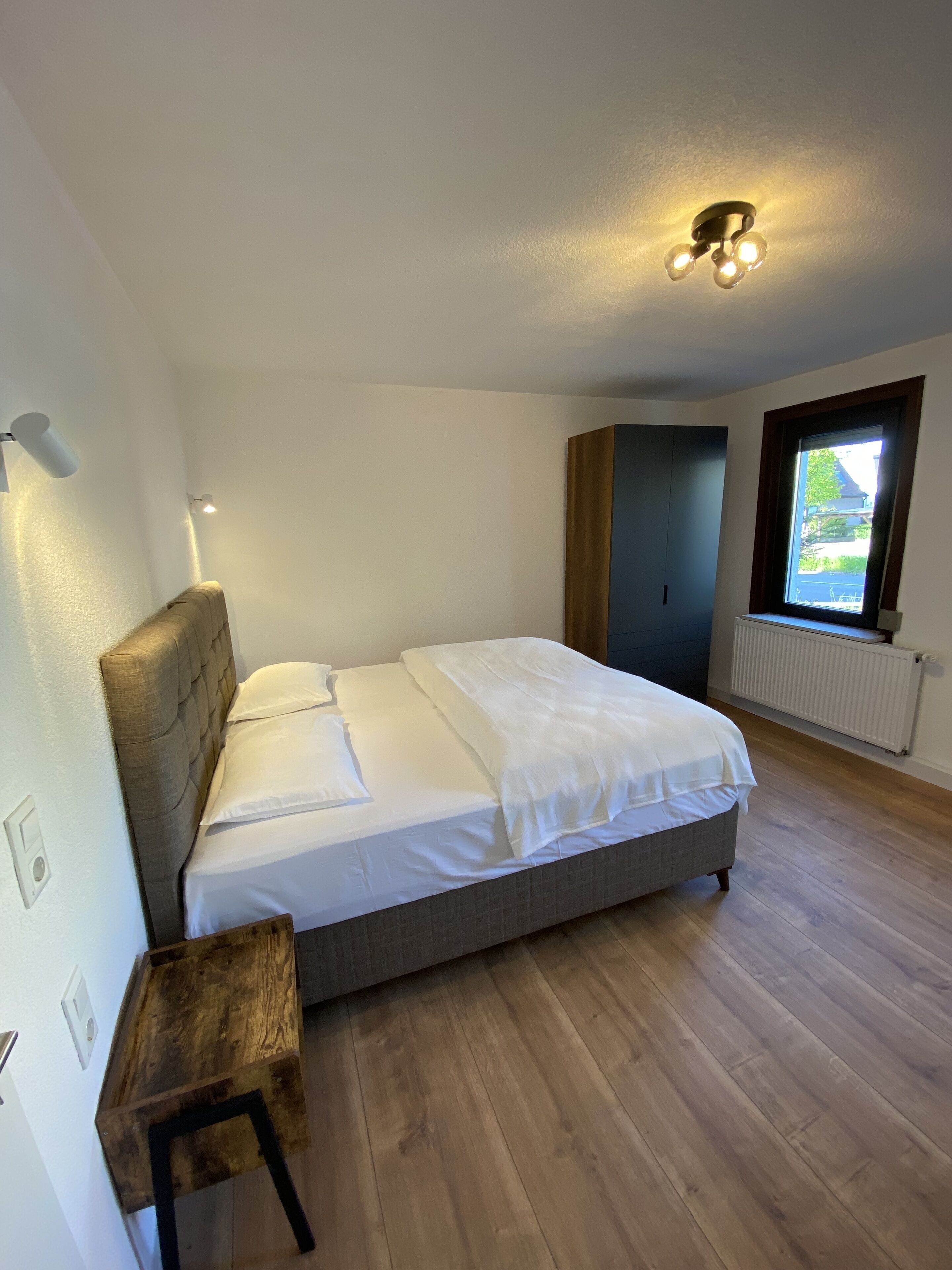 Ein Schlafzimmer mit Bett, einem Nachttisch aus Holz, einem Fenster mit Blick und einer Deckenleuchte.