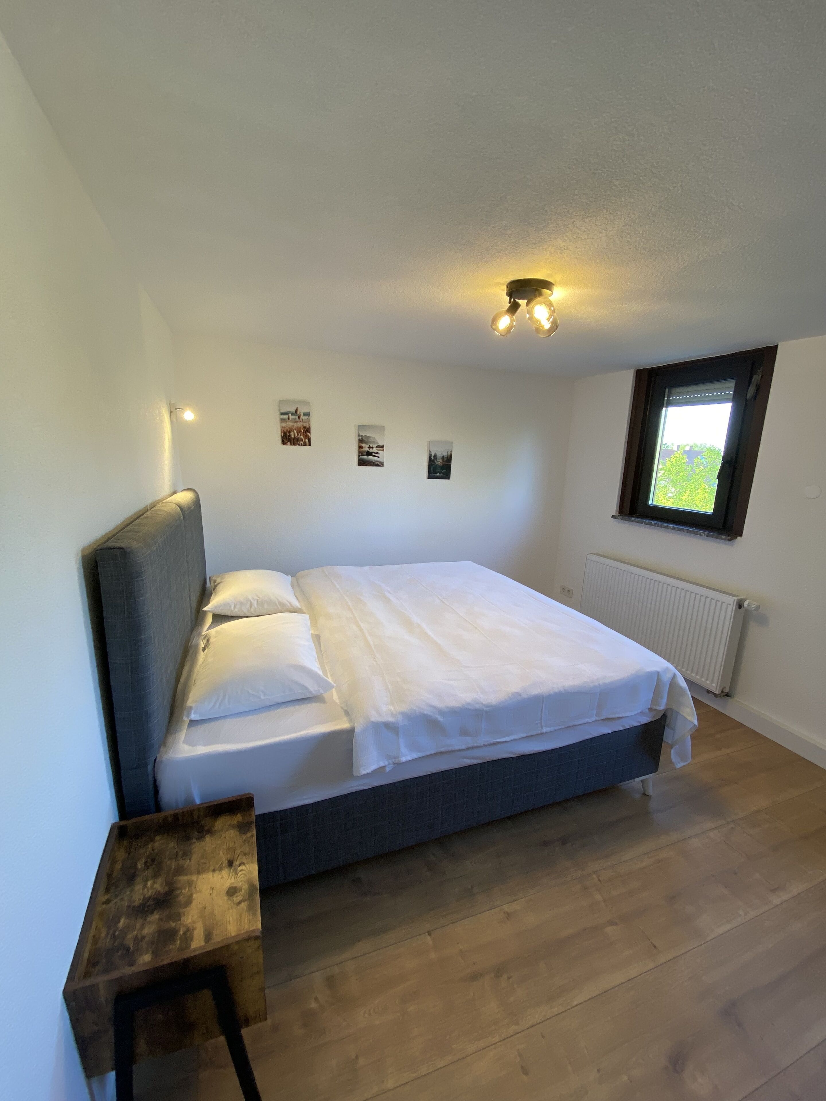 A bedroom with a bed, a small wooden table, a window, and wall-mounted lights.